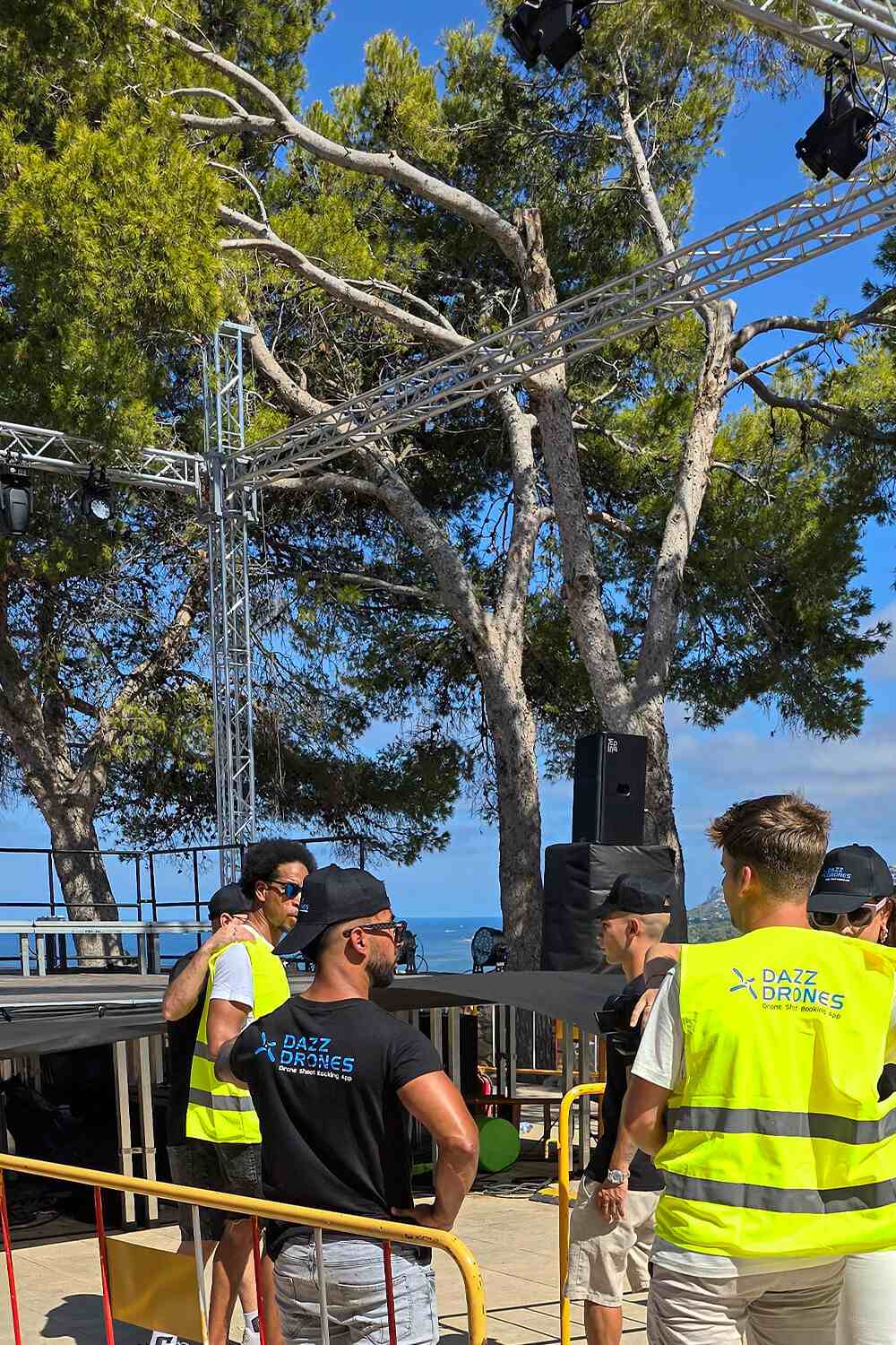 Dazz Drones pilot in branded cap and safety vest operating a drone remote control at an outdoor event, with tents and people visible in the background setting.