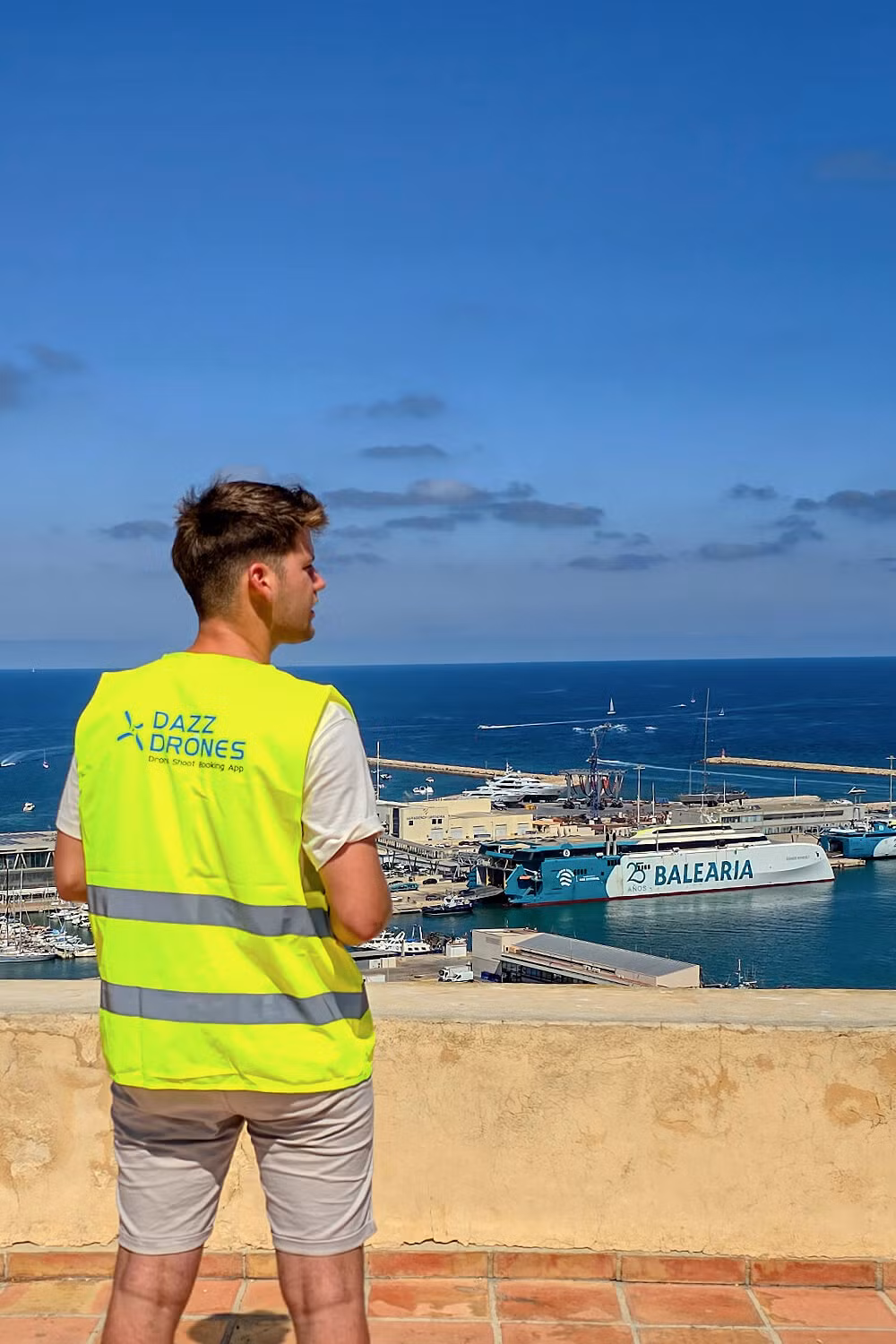 Dazz Drones pilot in safety vest overlooking a sunny harbor with boats and a Balearia ferry, scouting for a travel photography or videography project.