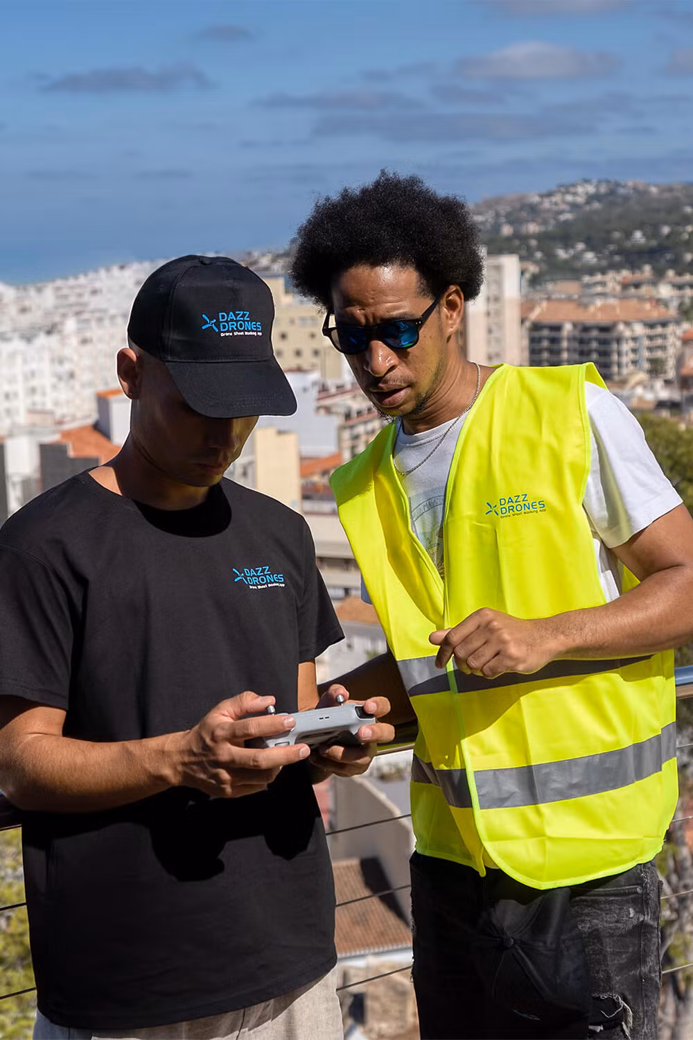 Two Dazz Drones pilots collaborating outdoors; one operates the drone remote while the other observes, conducting an aerial shoot for real estate with a city view.