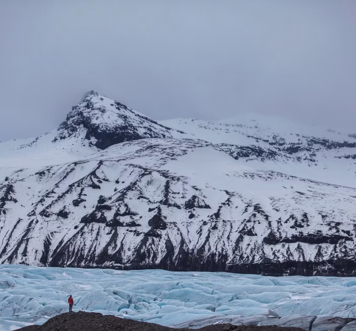 Vast Glacier Views in Skaftafell, Vatnajökull National Park, Iceland A small figure in a red jacket stands on a rocky outcrop overlooking the vast, crevassed ice field of a glacier in Skaftafell, Vatnajökull National Park, Iceland, with snow-dusted mountains behind under an overcast sky.