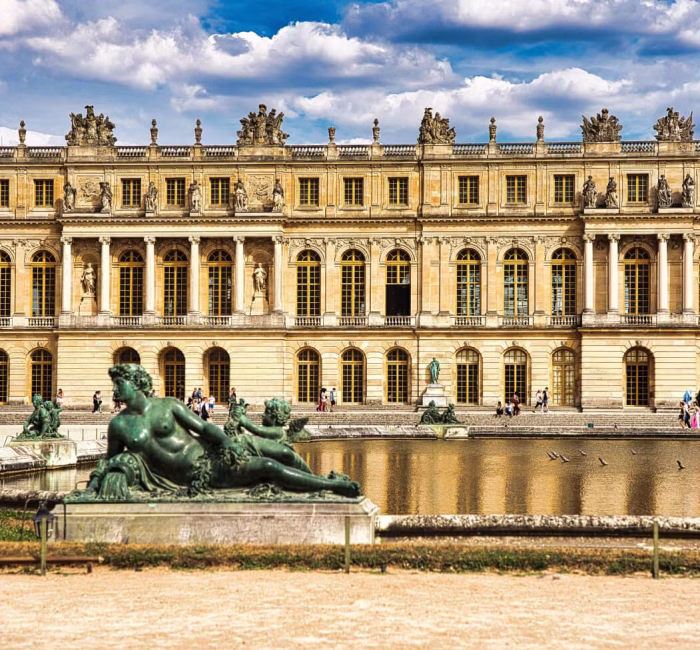 Palace of Versailles | Gardens View with Sculpture | France Exterior view of the Palace of Versailles in France, showing the grand building, a large water feature with classical sculptures, and visitors in the gardens.