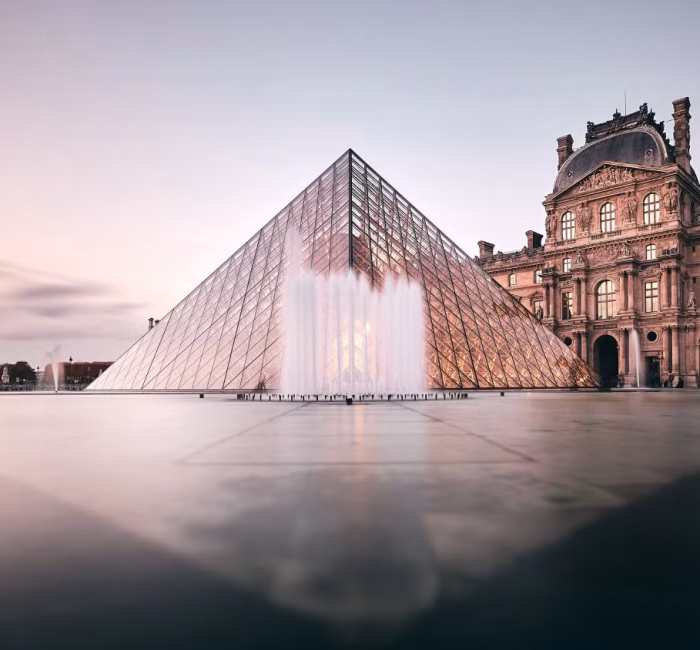 Louvre Museum Pyramid | Paris, France | Architectural Landmark The glass pyramid entrance of the Louvre Museum in Paris, France, with the historic palace building behind it, at dawn or dusk.