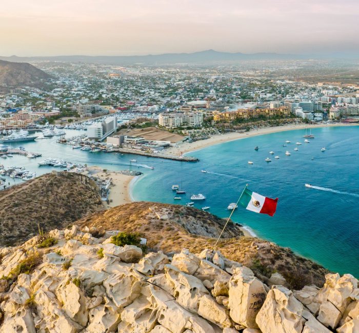 Vibrant flag waving in the foreground of a picturesque cityscape. Cabo San Lucas, Mexico. A vibrant flag waving in the foreground of a picturesque cityscape. Cabo San Lucas, Mexico.
